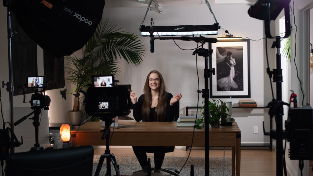 smiling brunette woman wearing black with hands up talking into a video production set up with camera and lighting equipment all around her 