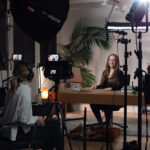 young brunette woman speaking into a camera by a young blonde woman who is performing video production for the brunette's business. She is sitting at a wooden table with lights and camera equipment around her